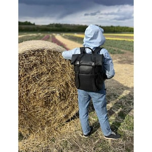 Peut inclure: Un sac à dos noir avec une finition texturée, doté d'une fermeture à enroulement et de sangles à boucle. Le sac à dos est porté par une personne en jean bleu et une veste bleu clair, près d'une botte de foin.
