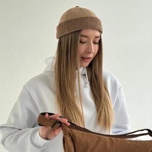 May include: A tan suede docker cap featuring a structured crown and a wide, ribbed corduroy cuff. The hat has a rounded silhouette with visible seam detailing along the top panels, providing a clean and uniform appearance for this headwear accessory.
