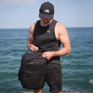 Op de afbeelding: Een zwarte rugzak met de tekst "USVIT" op de voorkant wordt vastgehouden door een persoon die een zwarte tanktop en korte broek draagt. De persoon draagt ook een zwarte baseballcap met witte tekst. De achtergrond is een watermassa.