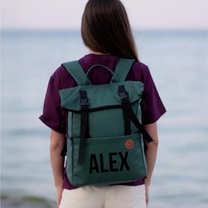 May include: A green backpack with black straps and buckles. The word "ALEX" is printed in large black letters on the front. A small brown circular patch is on the right side of the backpack.