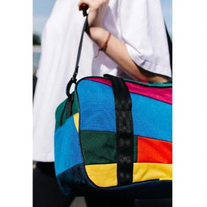 May include: A duffel bag featuring a vibrant patchwork design with rectangular panels in blue, green, yellow, red, and black. The bag includes black nylon webbing straps and black trim along the edges, constructed from a durable woven fabric.