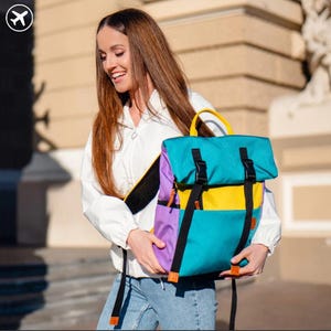 Könnte beinhalten: Ein farbenfroher Rucksack mit türkisfarbenen, gelben und lila Paneelen. Der Rucksack hat schwarze Riemen und Schnallen, einen gelben Griff und einen schwarzen Schultergurt. Die Tasche wird von einer Person getragen, die eine weiße Jacke und blaue Jeans trägt.