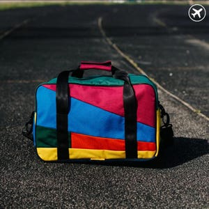 Op de afbeelding: Een rechthoekige sporttas met een kleurrijk ontwerp van rode, blauwe, groene en gele panelen. De tas is voorzien van twee zwarte stoffen handvatten, een bijpassende zwarte schouderbandbevestiging en een ritssluiting aan de bovenzijde.