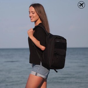 Op de afbeelding: Een zwarte rugzak met meerdere compartimenten en een gewatteerde rug wordt over de schouder gedragen. De rugzak heeft een rechthoekige vorm en een klein vakje aan de voorkant. De persoon draagt een zwart t-shirt en een spijkerbroek.