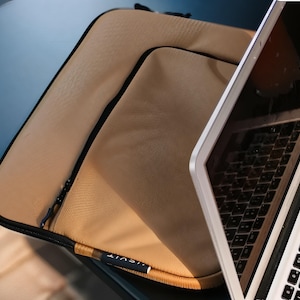 Könnte beinhalten: Eine beigefarbene Laptoptasche mit glatter, matter Textur und schwarzer Umrandung. Die Tasche verfügt über eine Vordertasche mit schwarzem Reißverschluss und ein kleines schwarzes Stoffetikett mit dem weißen Schriftzug "USVIT" an der Ecke.