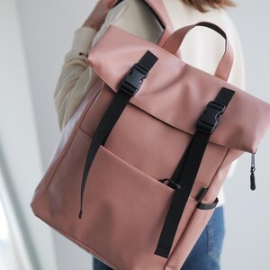 May include: A dusty rose backpack with black straps and buckles. The bag has a roll-top closure and a front pocket. The straps are adjustable, and the bag appears to be made of a textured material. A handle is visible at the top.