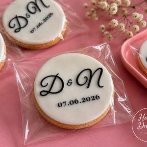 May include: Round sugar biscuits topped with smooth white icing, each individually wrapped in clear plastic. The biscuits feature the black printed text D & N and the date 07.06.2026 in a decorative script and sans-serif font combination.