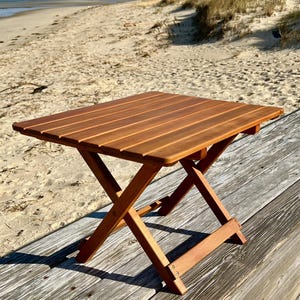 May include: A small, square, folding wooden table with a slatted top. The table is made of light brown wood and has an X-shaped leg design. The table is set up on a wooden deck near a sandy beach.