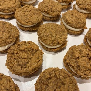 Puede incluir: Primer plano de varias galletas de crema de avena. Las galletas son redondas, con un exterior texturizado de color marrón dorado. Una gruesa capa de relleno de crema blanca es visible entre las dos mitades de la galleta. Las galletas están dispuestas sobre una superficie blanca.