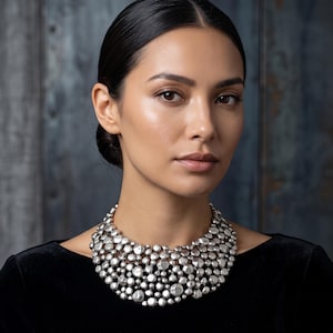 May include: A statement collar necklace featuring a dense cluster of polished, irregular silver-toned metallic spheres of varying sizes. The necklace sits high on the neck, creating a textured, reflective surface that covers the collarbone area.