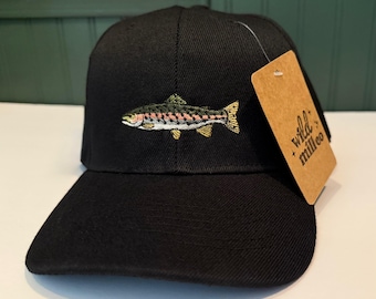 May include: Black baseball cap with an embroidered fish design. The fish is detailed with green, pink, and gold colors. A brown tag with the text "wild + milleo" hangs from the cap.