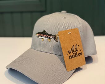 May include: A gray baseball cap with an embroidered fish design. The cap has a curved brim and a brown tag that reads "wild mill co". The fish is detailed with shades of brown, pink, yellow, and black.