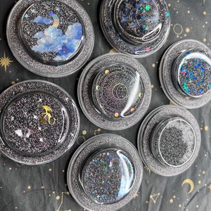 Op de afbeelding: Een verzameling van zeven ronde, heldere hars onderzetters met glitter en hemelse ontwerpen. Elke onderzetter heeft een ander ontwerp, waaronder wolken, manen, sterren en een draak. De onderzetters hebben een zilveren glitterrand.