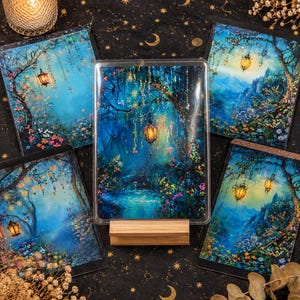Könnte beinhalten: Eine Sammlung von Fantasy-Kunstdrucken, die eine mystische Waldszene mit leuchtenden Laternen, Blumen und einem ruhigen Teich zeigen. Die Drucke sind auf schwarzem Stoff mit goldenen Himmelsmustern und einer brennenden Kerze angeordnet.