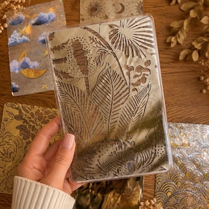 Könnte beinhalten: Eine Hand hält eine transparente Kunststoffhülle mit einem detaillierten botanischen Design. Mehrere dekorative Karten mit Himmels- und Blumenmustern sind auf einer Holzoberfläche angeordnet. Die Karten zeigen Sonnen, Monde, Wolken und Blättermuster.