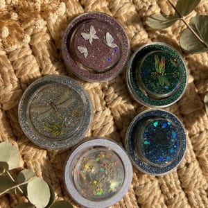 Könnte beinhalten: Fünf kreisförmige Untersetzer aus Kunstharz mit eingearbeitetem Glitzer und Naturmotiven. Einer zeigt weiße Schmetterlinge, ein anderer eine grüne Libelle, ein dritter eine silberne Libelle, ein vierter eine blaue Libelle und der letzte goldene Schmetterlingsumrisse.