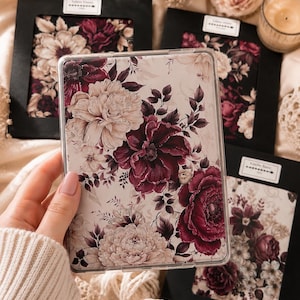 Könnte beinhalten: Eine transparente E-Reader-Hülle mit einem Blumenmuster aus großen cremefarbenen und dunkelburgunderroten Blüten auf hellem Grund. Das Design zeigt detaillierte dunkle Blätter und Stängel und bietet eine botanische Ästhetik auf der gesamten Oberfläche.
