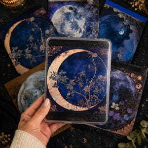 Könnte beinhalten: Eine transparente Handyhülle mit einem Halbmond-Design und floralen Akzenten in Gold und Marineblau. Die Hülle wird in einer Hand gehalten, umgeben von Schreibwaren mit ähnlichen Himmelsdesigns. Eine brennende Kerze befindet sich im Hintergrund.