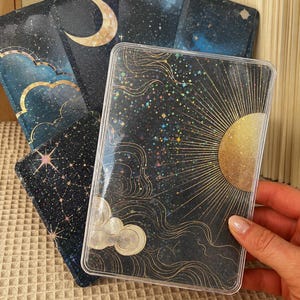 Könnte beinhalten: Ein Set Tarotkarten mit Himmelsmotiven. Die Karten haben einen dunkelblauen Hintergrund mit goldenen Akzenten, darunter ein Halbmond, eine Sonne, Sterne und Wolken. Eine transparente Kunststoffhülle schützt die Karten. Die Karten werden von einer Person gehalten.
