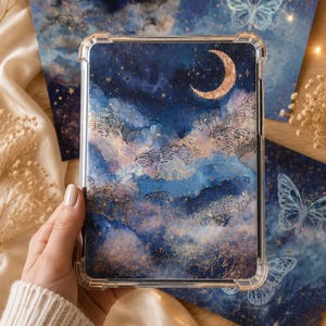 Könnte beinhalten: Eine transparente Handyhülle mit einem Himmelsdesign, das einen Mond, Sterne und Wolken in Blau-, Gold- und Weißtönen zeigt. Passende Kunstdrucke sind im Hintergrund zu sehen.