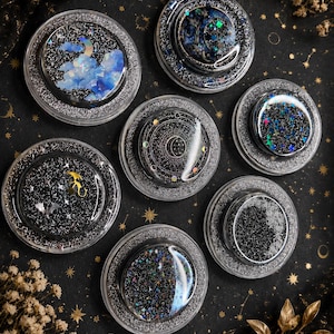 May include: A collection of seven round, clear resin coasters with celestial designs. Each coaster features a different cosmic scene with glitter, stars, and moon accents. The coasters are set against a black background with gold star and constellation patterns.