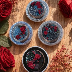 Könnte beinhalten: Vier runde Behälter mit floralen Designs, die rote Rosen und grüne Blätter zeigen, werden auf einer Holzoberfläche präsentiert. Die Behälter haben silberne Glitzerkanten und unterschiedliche Hintergründe in Blau und Schwarz. Rote Rosen und Blätter sind verstreut.
