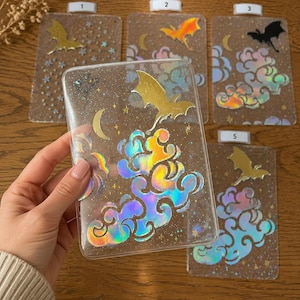 Könnte beinhalten: Transparente Handyhüllen mit verschiedenen Designs. Eine Hülle zeigt eine goldene Fledermaus, schillernde Wolken und einen Mond. Andere Hüllen zeigen Fledermäuse in Gold, Orange und Schwarz, mit Sternen und Wolken. Die Hüllen sind rechteckig und haben einen Glitzereffekt.