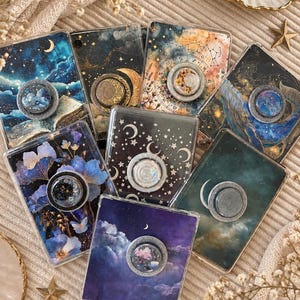 Könnte beinhalten: Eine Sammlung von acht rechteckigen Handyhüllen, jede mit einem einzigartigen Himmels- oder Blumenmotiv und einem runden, einklappbaren Griff in der Mitte. Die Designs zeigen Sternenhimmel, Sternbilder, Monde, Wolken und blaue Blumen.