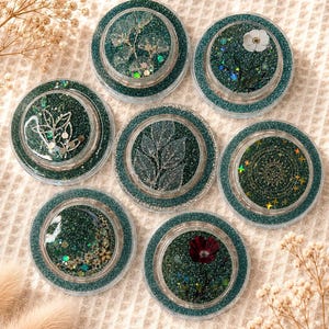Könnte beinhalten: Sieben runde Untersetzer mit einem tealfarbenen Glitzerrahmen und einem dunkelgrünen Mittelteil. Jeder Untersetzer hat ein anderes Design, darunter getrocknete Blumen, Blätter und ein Sonnenmuster. Die Untersetzer sind auf einer cremefarbenen, strukturierten Oberfläche angeordnet.