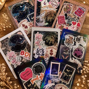 Könnte beinhalten: Eine Sammlung dekorativer, harzbeschichteter Buchcover mit Glitzerkanten. Jedes Cover zeigt Aufkleber mit himmlischen, floralen und gotischen Themen, darunter Texte wie "Hot Girls Read", "Dark Romance Book Club" und "Enemies to Lovers."