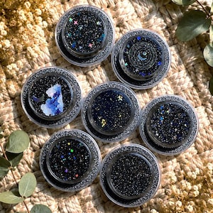 Könnte beinhalten: Sieben kreisförmige Behälter gefüllt mit dunklem, glitzerndem Harz. Jeder Behälter zeigt ein einzigartiges himmlisches Design mit holografischen Sternen, Mondsicheln und wolkenartigen Mustern in tiefen Blau-, Schwarz- und schimmernden Silbertönen.