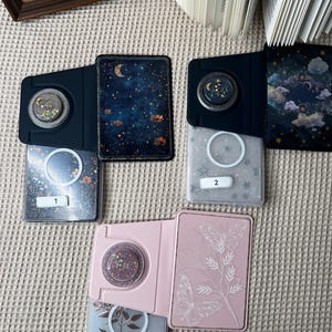 May include: A collection of phone grip accessories with various designs. The grips feature glitter, celestial, and floral patterns. The cases are in shades of pink, blue, and black. Some cases have the numbers 1, 2, and 3.
