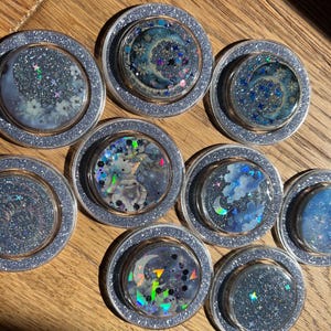 Può includere: Una collezione di sottobicchieri rotondi e glitterati con disegni celesti. Ogni sottobicchiere presenta un centro trasparente e circolare con un design unico di lune, stelle e nuvole, circondato da un bordo glitterato argentato. Esposti su una superficie di legno.