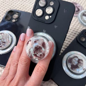 Op de afbeelding: Zwarte telefoonhoesjes met de tekst 'Pro Camera' en hartpatronen. Eén hoesje heeft een popsocket met een hondengezicht, een ander een foto van twee mensen. Andere hoesjes met vergelijkbare popsockets zijn ook zichtbaar.
