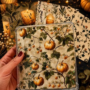 Könnte beinhalten: Eine transparente Tablet-Schutzhülle mit einem Druckmuster aus orangefarbenen Kürbissen, grünen Blättern und kleinen Beeren auf cremefarbenem Hintergrund. Das Design bedeckt die gesamte Rückseite der rechteckigen Hülle mit verstärkten Ecken.