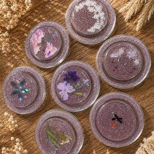 Könnte beinhalten: Eine Sammlung von sieben runden Untersetzern mit glitzernder lila Basis und klaren, erhöhten Rändern. Jeder Untersetzer hat ein einzigartiges Design, darunter gepresste Blumen und florale Muster. Die Untersetzer sind auf einer Holzoberfläche angeordnet.
