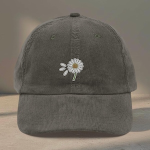 May include: Olive green corduroy baseball cap with a white daisy embroidered on the front. The daisy has a yellow center and is accompanied by two white petal accents. The cap has a curved brim and a rounded top.