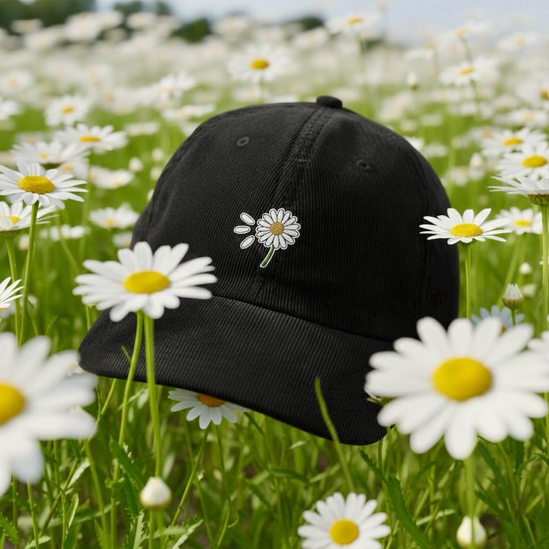May include: Black baseball cap with a daisy flower patch. The cap is set in a field of daisies with white petals and yellow centers. The hat has a curved brim and a small button on top.