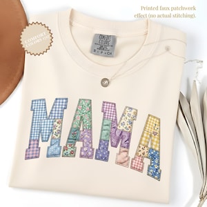 Puede incluir: Camiseta crema de cuello redondo con la palabra "MAMA" estampada en la parte delantera con un diseño de patchwork. Las letras de patchwork presentan varias telas florales y estampadas. Un collar plateado con un colgante está sobre la camiseta.
