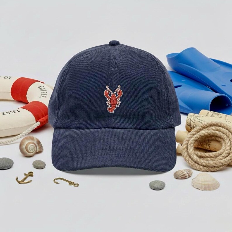 May include: Navy blue corduroy baseball cap with a red lobster embroidered on the front. The cap has a curved brim and a rounded crown. The background includes nautical-themed items like a life preserver, seashells, and blue flippers.