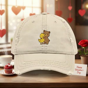 Pode incluir: Boné de beisebol creme desgastado com um design bordado de um urso marrom abraçando um pato amarelo. O boné tem uma viseira curva e uma aparência desgastada. O design está centrado na frente. Um cartão diz "Happy Valentine's Day".