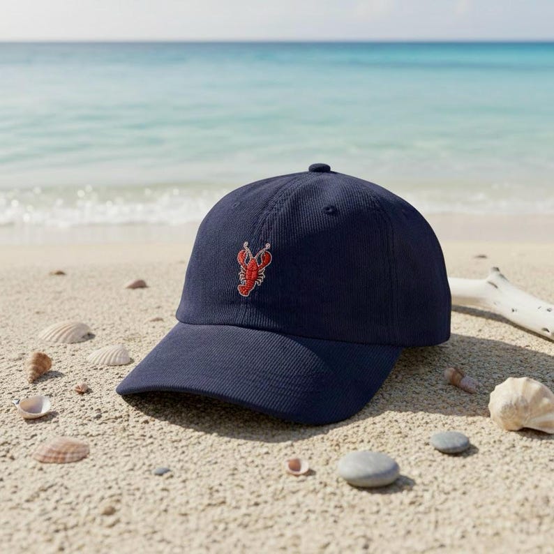 Puede incluir: Gorra de b&eacute;isbol azul marino con un dise&ntilde;o de langosta roja bordada. La gorra est&aacute; hecha de una tela similar a la pana y tiene una visera curva. La gorra est&aacute; sobre una playa de arena con conchas marinas.