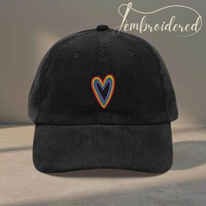 May include: Black corduroy baseball cap with a rainbow heart embroidered on the front. The cap has a curved brim and a top button. The word "embroidered" is written in cursive on the top right of the cap.