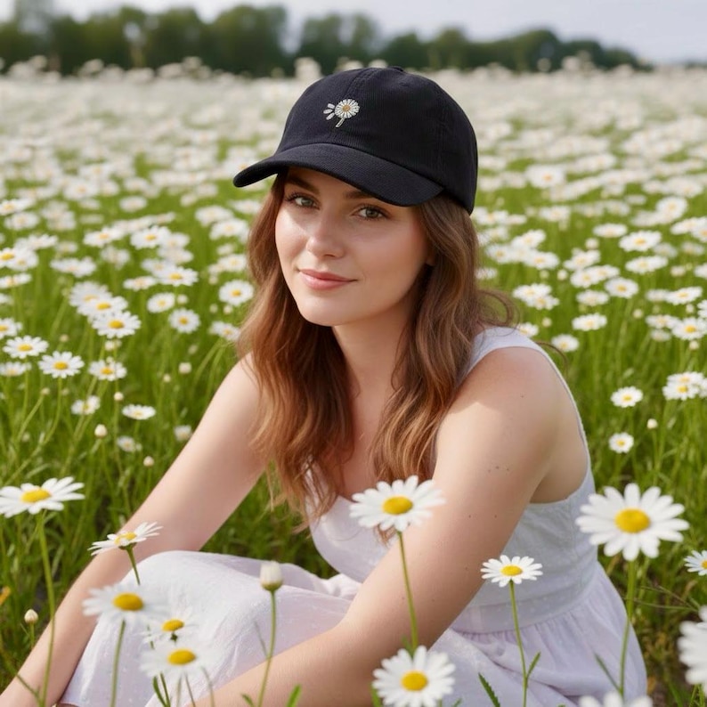 May include: A black baseball cap with a small embroidered daisy on the front. The cap is worn by a person sitting in a field of daisies. The person is wearing a white dress.
