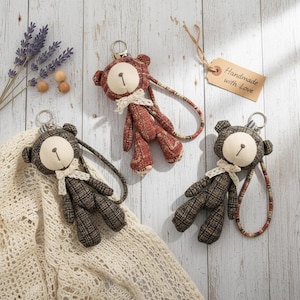 May include: Three handmade teddy bear keyrings in brown and red hues. Each bear features a cream-coloured face, a small bow, and a long strap. One keyring has a tag that says "Handmade with Love".