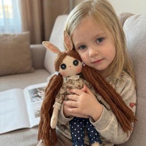 May include: A handmade doll with long brown hair, large black button eyes, and bunny ears. The doll wears a floral print dress and blue polka dot pants. The doll is being held by a young girl.