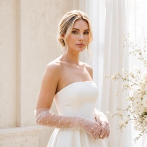 Può includere: Un abito bianco senza spalline abbinato a guanti in tulle trasparente lunghi fino al gomito. I guanti sono realizzati in un tessuto a rete fine e traslucido, decorati con piccole perle bianche distribuite uniformemente lungo tutta la manica.