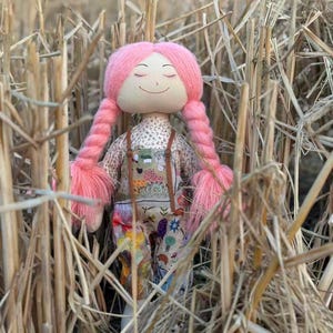 Op de afbeelding: Een handgemaakte pop met een lachend gezicht en gesloten ogen, met lange roze gevlochten haar. De pop draagt een top met bloemenpatroon en een overall met kleurrijke ontwerpen. De pop staat tegen een achtergrond van droog gras.