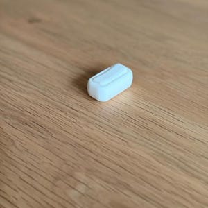 Op de afbeelding: Een klein, wit, rechthoekig object met een gebogen bovenkant, rustend op een houten oppervlak. Het object lijkt een kleine container of een onderdeel van een groter item te zijn. De achtergrond is een lichtbruine houtnerf.