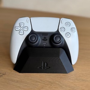Op de afbeelding: Een wit en zwarte PlayStation 5-controller op een zwart oplaadstandaard. De controller heeft een symmetrisch ontwerp met knoppen en joysticks. De standaard heeft het PlayStation-logo.